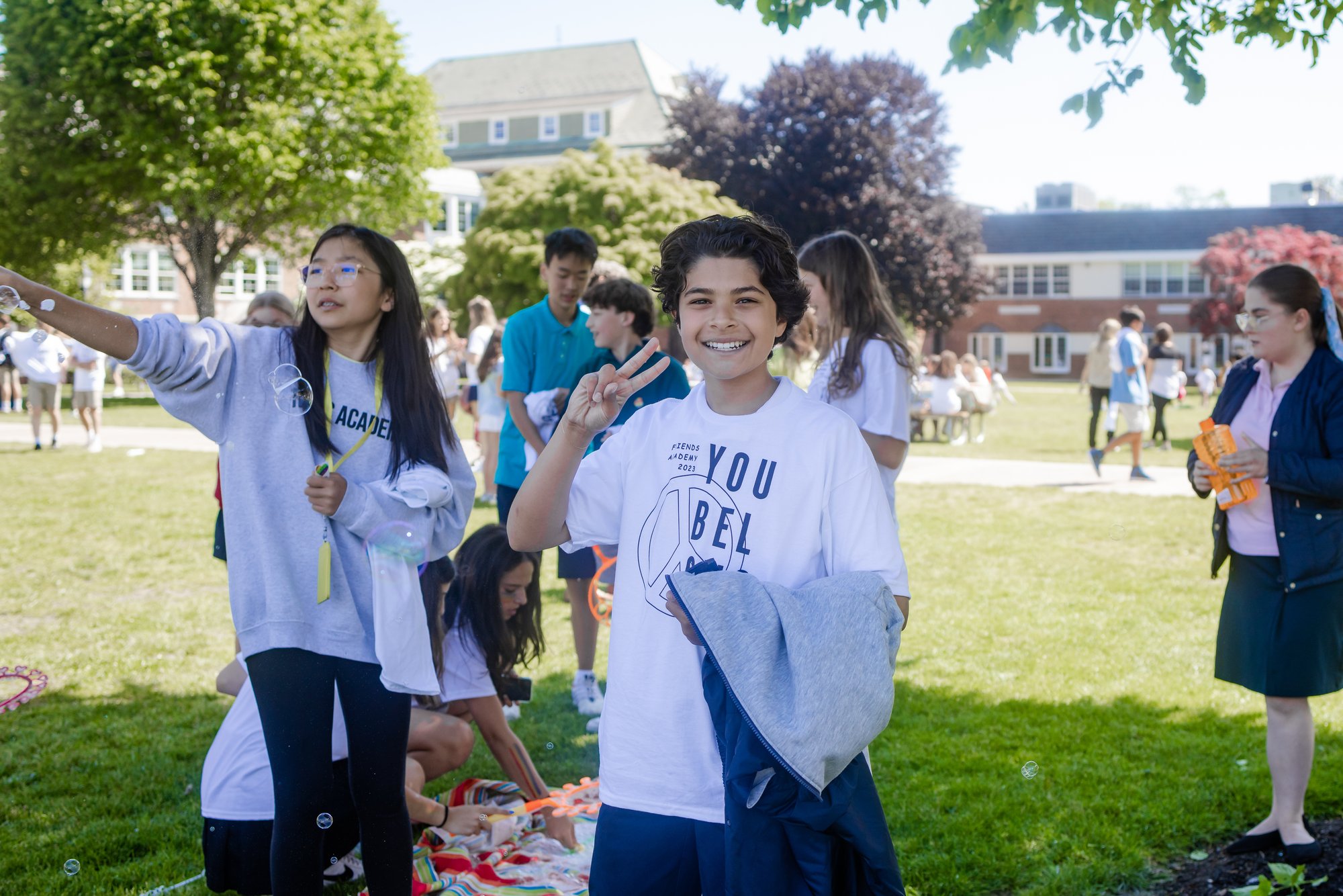 A boy smiles and holds up a peace sign at an outdoor event on a sunny day. People in the background engage in activities like blowing bubbles. 