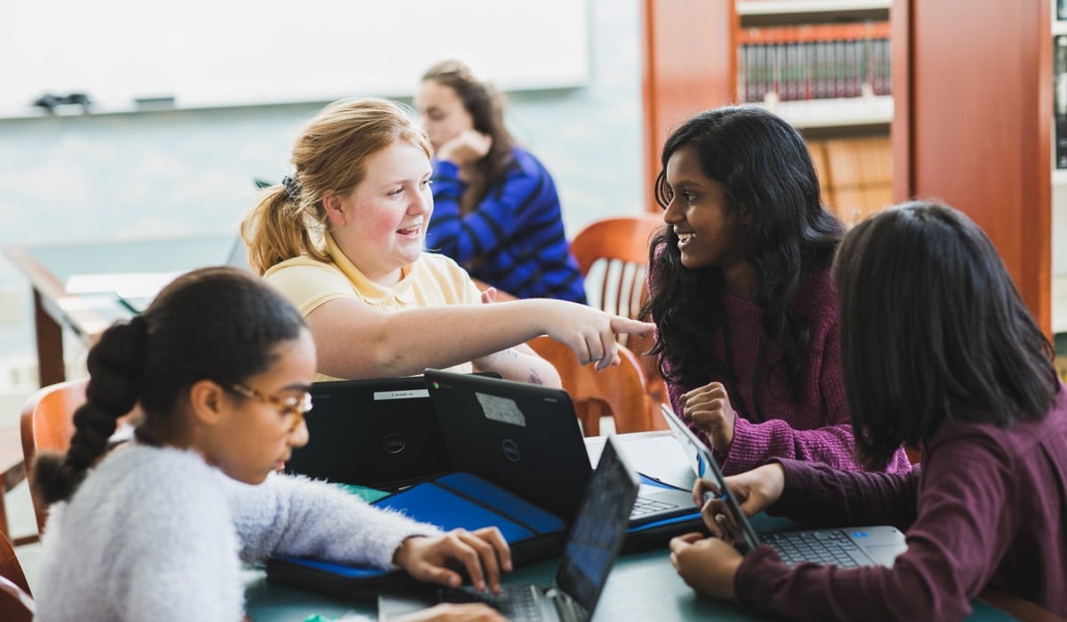 Four students seated at a table, working on laptops and tablets, in a classroom setting. One student is pointing at another's screen. Bookshelves and a whiteboard are in the background.